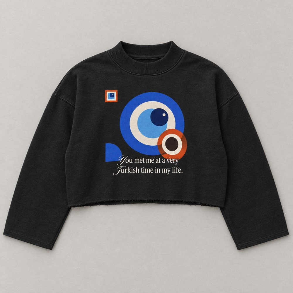 AI visualisation: black boxy sweatshirt with mock neck and raw-edge hem; front print with a Bauhaus-style nazar and the line “You met me at a very Turkish time in my life.”