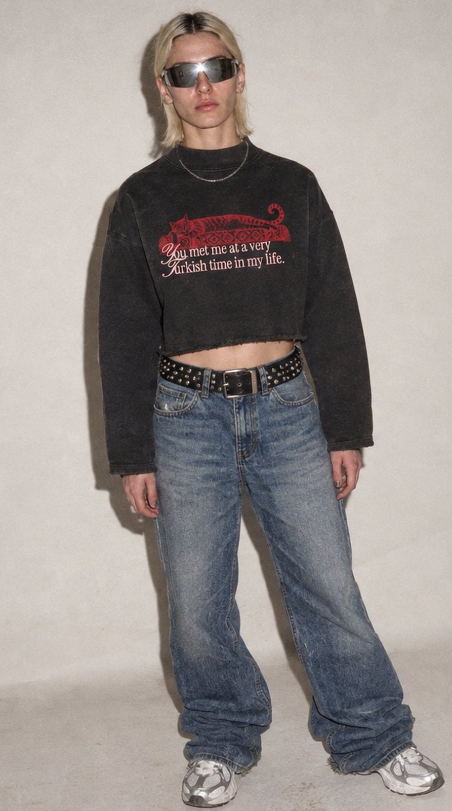 Frau mit Sonnenbrille vor grauer Studiowand, im schwarzen Kedi-Sweatshirt, Baggy-Jeans