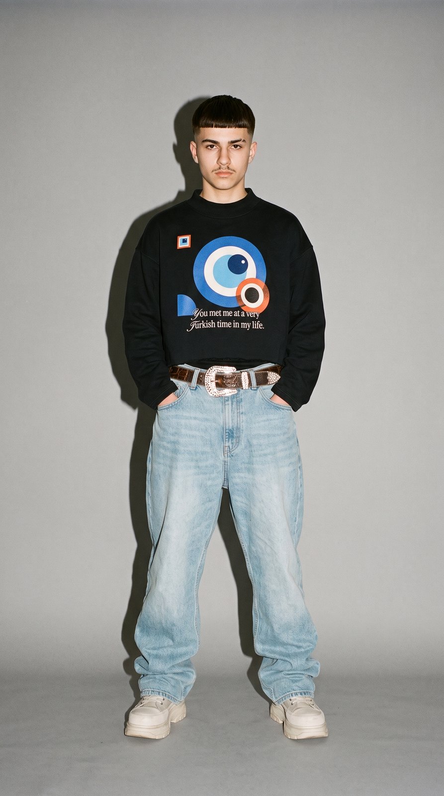 Junger Mann im Studio mit Direktblitz, im schwarzen Boxy Sweatshirt A Very Turkish Time mit Bauhaus-Nazar-Print, helle Baggy-Jeans