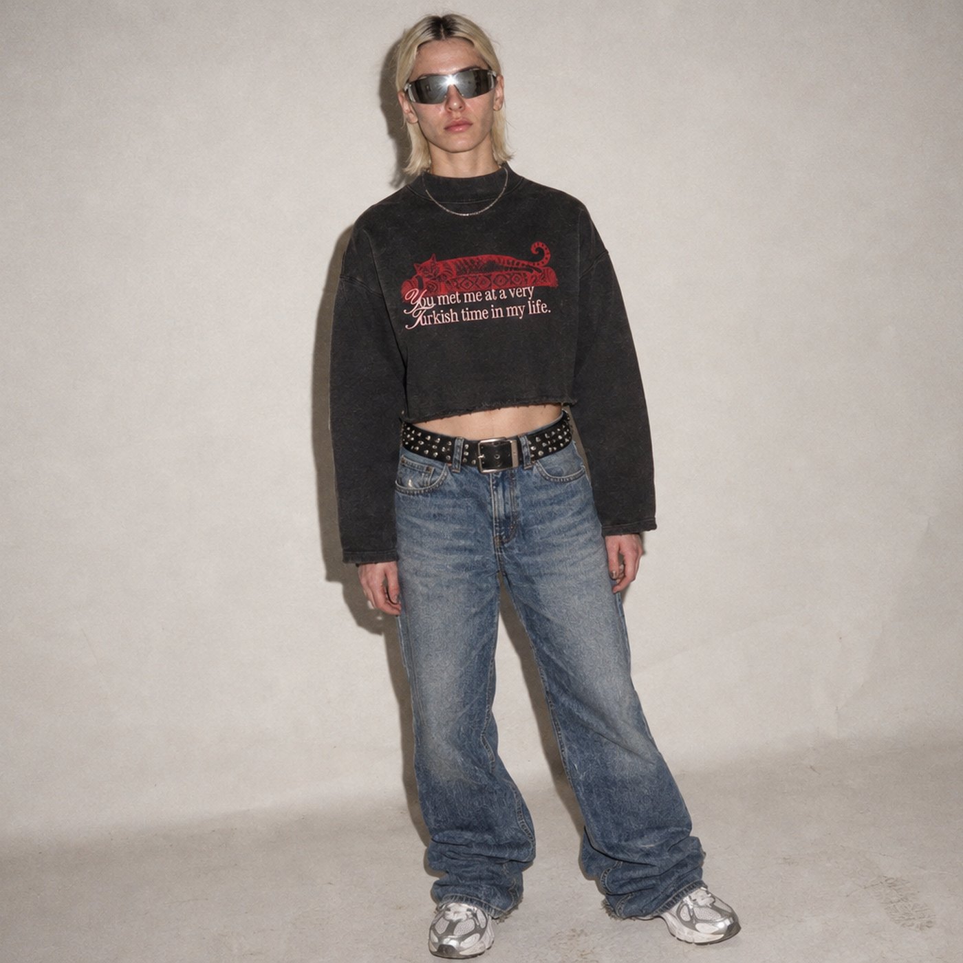 Frau mit Sonnenbrille vor grauer Studiowand, im schwarzen Kedi-Sweatshirt, Baggy-Jeans