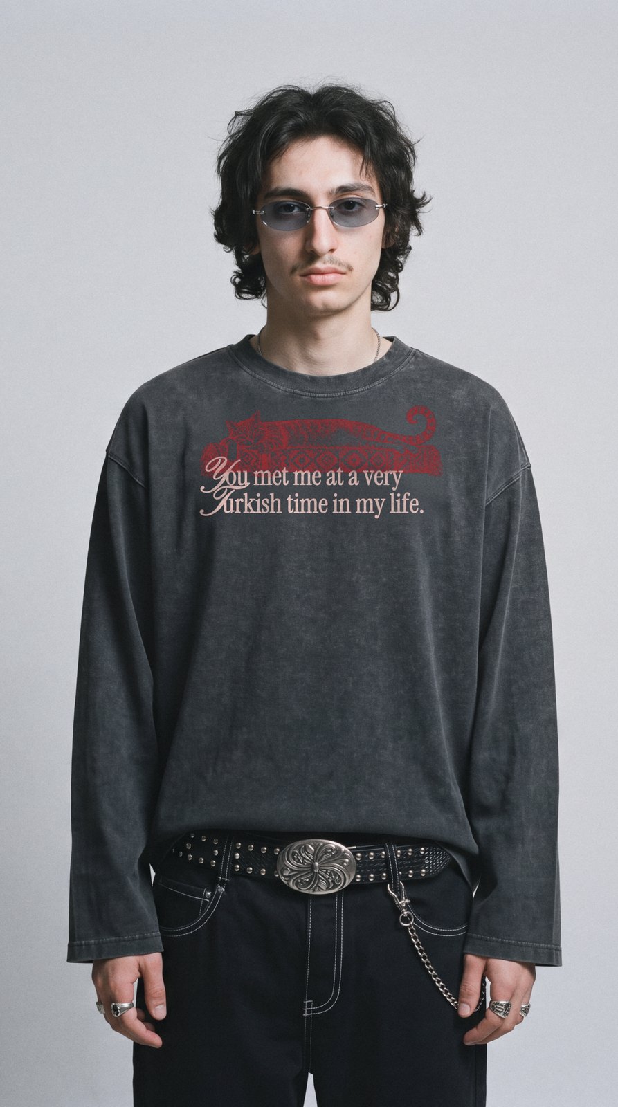 Junger Mann mit ovaler Sonnenbrille vor grauer Studiowand, im sun-faded schwarzen Kedi-Longsleeve, Saum am Gürtel