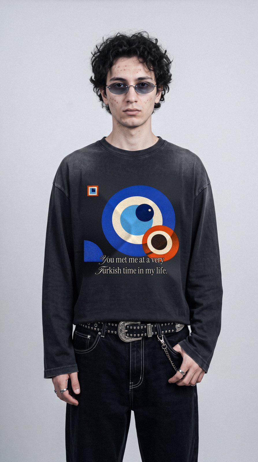 Junger Mann mit Locken und ovaler Sonnenbrille vor grauer Studiowand, im sun-faded schwarzen Longsleeve A Very Turkish Time mit Bauhaus-Nazar-Print, Saum am Gürtel