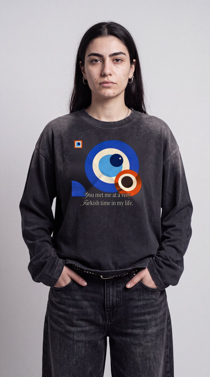 Junge Frau mit Creolen vor grauer Studiowand, im sun-faded schwarzen Longsleeve mit Nazar-Print, Hände in den Taschen