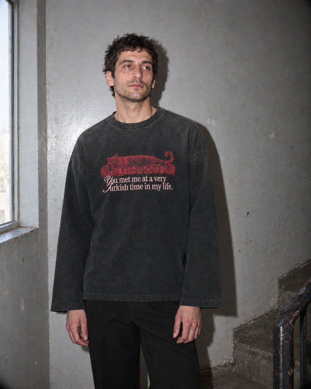 Junger Mann im Treppenhaus, im sun-faded schwarzen Kedi-Longsleeve mit roter Katze auf Kelim