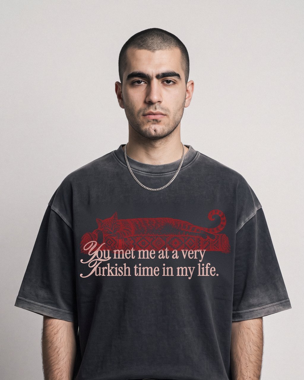 Junger Mann mit Buzzcut vor grauer Studiowand, im sun-faded schwarzen Boxy T-Shirt mit roter Katze auf Kelim, Saum über dem Gürtel