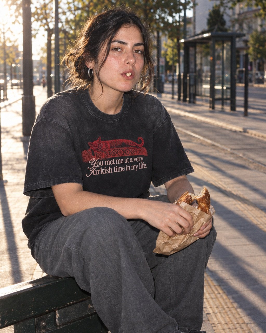 Junge Frau sitzt im Abendlicht an einer Haltestelle, im sun-faded schwarzen Kedi-T-Shirt, mit Simit in der Hand
