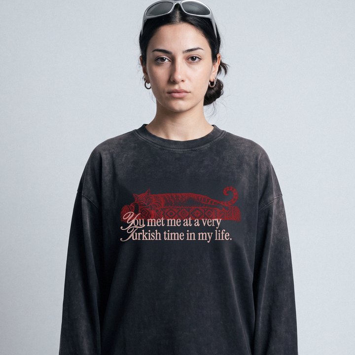 Junge Frau vor grauer Studiowand im sun-faded schwarzen Kedi-Longsleeve mit roter Katze auf Kelim
